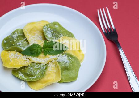 Teller mit köstlichen grünen und gelben Ravioli mit Käse und Basilikum auf rotem Hintergrund. Italienisches Essen. Stockfoto