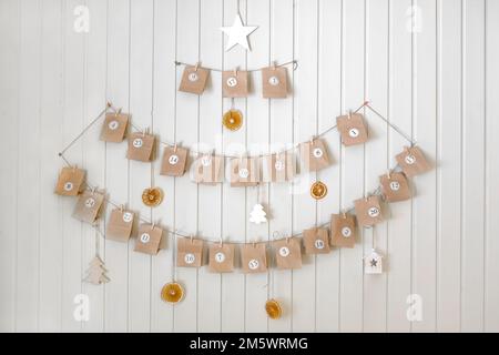 Adventskalender. Kleine handgemachte Papiergeschenke hängen auf dem Hintergrund einer Holzwand, Nahaufnahme. Weihnachtskalender, Geschenk für Kinder. HEIMWERKER. Sustainabl Stockfoto