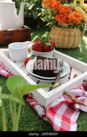 Ein Tablett mit einem Stück Schokoladenkuchen auf einem weißen Teller, einer Tasse Tee und einer Schüssel Erdbeeren auf einem Blumenhintergrund in einer Rattanvase Stockfoto