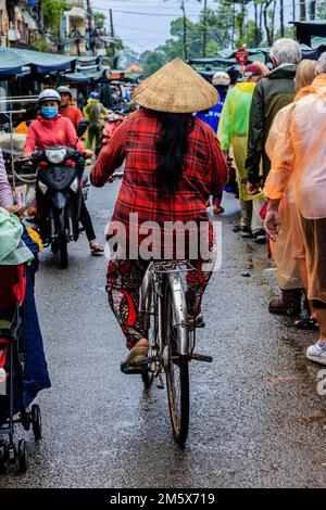 vietnamesische Dame in traditionellem roten Outfit und konischem Hut fährt sich durch den überfüllten Markt in Sa Dec Vietnam Stockfoto
