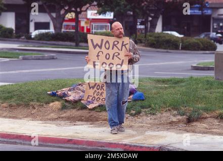 Ein Obdachloser steht am Straßenrand und hält ein Schild mit der Aufschrift Arbeit für Essen Stockfoto