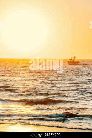 Eine Vertikale eines entfernten Schiffs auf einer Reise in der welligen Meereslandschaft vor dem glühenden Sonnenuntergangshimmel Stockfoto