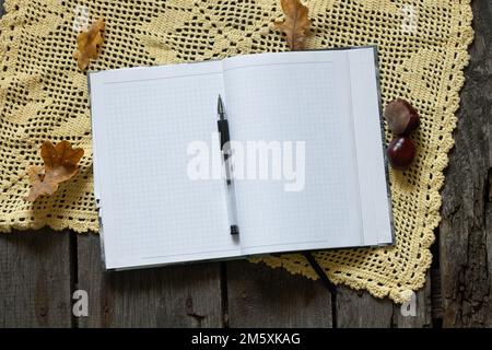 Kariertes Notizbuch mit sauberen flachen Karten und Stift auf Holztisch und Herbstblättern mit Kastanien, Arbeitsplatz, Herbstmotiv Stockfoto