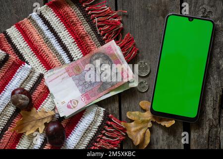 Telefon mit grünem Bildschirm und ukrainischer Hrywna und Münzen auf einem Holztisch mit Eichenblättern und Kastanien, Herbstthema, Online-Bezahlung Stockfoto