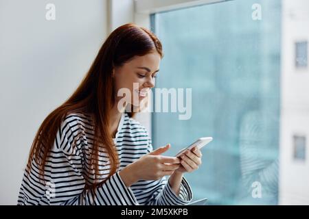 Frau mit einem Lächeln am Telefon, mit Zähnen vor einem Fenster mit Blick auf die Stadt, das Konzept des Online-Arbeitens und -Lernens über das Internet Stockfoto