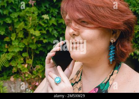 Profilporträt einer jungen Kaukasierin, rothaarig plus Model, die am Telefon telefoniert und aufmerksam zuhört, während sie im Park wartet. Stockfoto