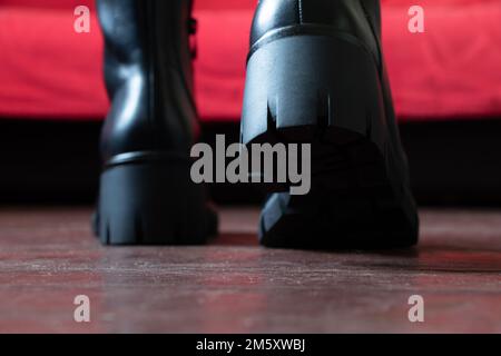 Mädchen in hohen schwarzen Stiefeln auf dem alten schmutzigen Boden zu Hause, Damenschuhe, Füße in Schuhen auf dem Boden, Schuhe auf dem Holzboden Stockfoto