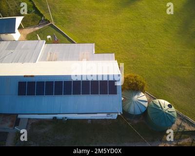 Sonnenkollektoren aus der Vogelperspektive auf einem Gebäudedach mit grünem Rasen, Spielplatz, Wassertanks und Außengebäude. Victoria, Australien. Stockfoto