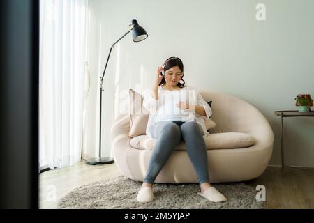 Porträt einer schwangeren Happy Asian Woman, die zu Hause auf dem Sofa sitzt und ein Smartphone benutzt, um Musik über Kopfhörer zu hören. Stockfoto