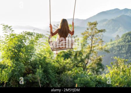Ein Mädchen, das auf dem Schaukelseil sitzt, hängt von der Schaukel des Astes und bietet einen wunderschönen Blick auf die Berge Stockfoto