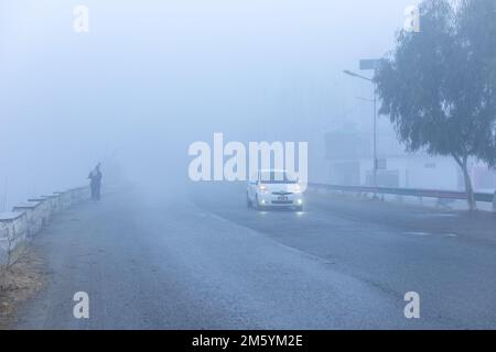 Ein Mann, der früh am Morgen in starkem Nebel auf der Straße unterwegs ist, während das Auto auf einer Straße mit schlechter Sicht unterwegs ist Stockfoto