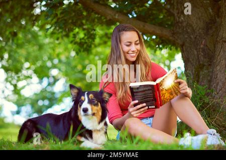 Im Park mit ihrem besten Kumpel chillen. Eine junge Frau, die ein Buch liest, während sie mit ihrem Hund unter einem Baum sitzt. Stockfoto