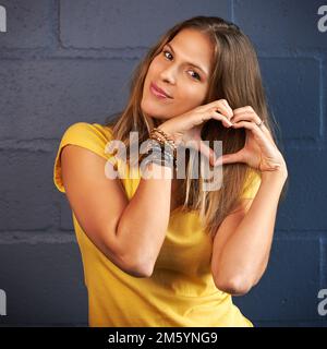 Liebe Grüße von mir an dich. Porträt einer jungen Frau, die eine Herzgeste macht, mit den Händen vor einem Backstein-Hintergrund. Stockfoto