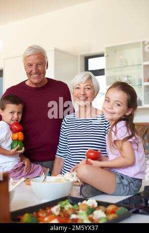 Es ist immer ein toller Tag mit Oma und Opa. Porträt von Großeltern, die mit ihren Enkelkindern in der Küche Abendessen zubereiten. Stockfoto