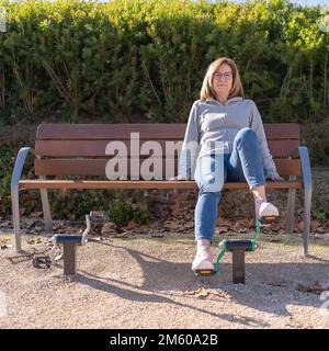 Reife Frau, die auf einer Bank sitzt und in einem öffentlichen Park auf einem Gymnastikgerät in die Pedale tritt. Stockfoto