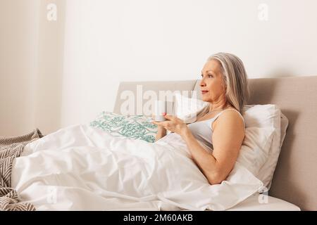Seniorin mit einem Becher. Alte Frau am Morgen im Schlafzimmer. Stockfoto