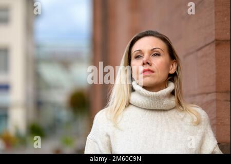 Eine attraktive blonde Frau mittleren Alters steht vor einer alten braunen Kirche in der Stadt und schaut aufmerksam in die Ferne Stockfoto