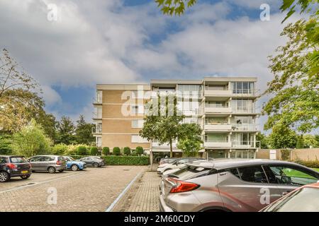 Ein Parkplatz mit Autos, die an einem wolkigen, blau-weißen Tag vor einem Wohnhaus geparkt sind Stockfoto