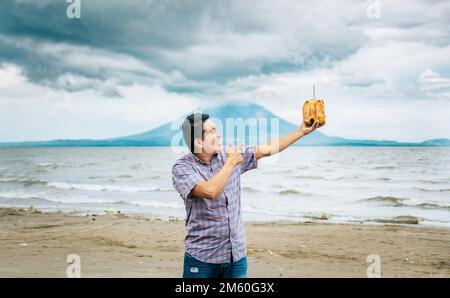 Junger Mann, der Urlaub macht und eine Kokosnuss am Strand genießt. Ein lächelnder Mann, der eine Kokosnuss hält und auf einen Strand in Nicaragua zeigt. Junger Mann zeigt Stockfoto