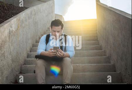 Gutaussehender Mann, der auf Treppen sitzt und sein Smartphone benutzt, Vorderansicht eines Mannes, der auf Treppen sitzt und sein Handy draußen benutzt, Nahaufnahme eines jungen Mannes, der darauf sitzt Stockfoto