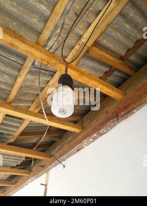 Nahaufnahme einer weißen Lampe und von einem Holzrahmen unter einem Blechdach hängende Drähte. Indonesien. Ansicht aus niedrigem Winkel Stockfoto