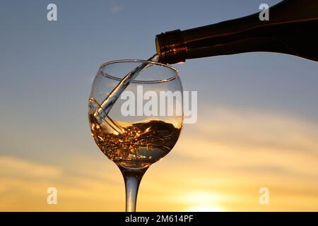 Weißwein, der aus einer Flasche in das Glas strömt, vor dem Hintergrund des Sonnenuntergangs am Himmel. Feier, romantisches Abendessen mit alkoholischen Getränken Stockfoto