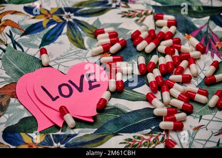 Rosa Herzen in einem von ihnen schrieben das Wort Liebe mit einem Haufen Pillen, auf einem Tisch mit einem tropischen Muster. Stockfoto