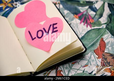 Öffnen Sie das Notizbuch zu einem leeren Blatt mit zwei rosa Herzen auf einem Blatt, das das Wort „Love“ auf einem Tisch mit einem tropischen Muster schrieb. Stockfoto