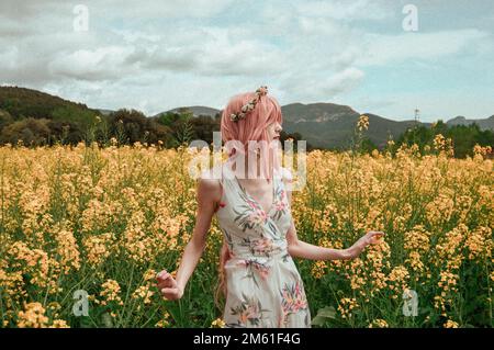 Mädchen in einem langen Sommerkleid, trägt eine rosa Perücke und eine Blumenkrone, Bilder auf einem Blumenfeld, mit klarem Himmel und traumhaftem Filter Stockfoto