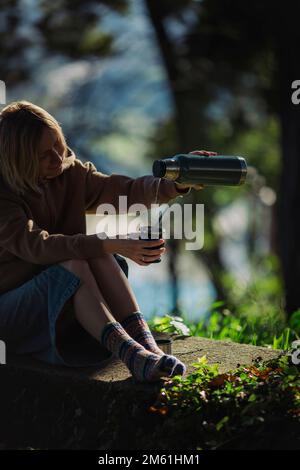 Eine Frau, die im Park sitzt, gießt heißes Wasser aus einer Thermoskanne in einen Mate Becher. Stockfoto