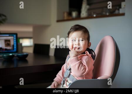 Neun Monate altes süßes Baby, das in einem pinkfarbenen Babystuhl sitzt, lächelt und in den Finger beißt. Stockfoto