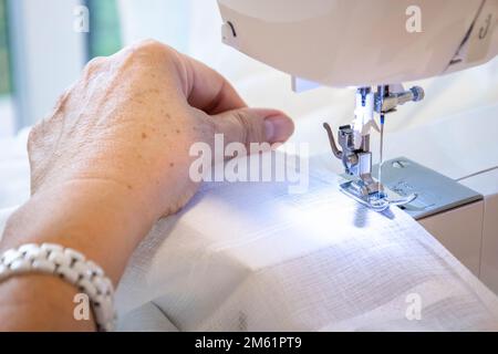 Nahaufnahme der oberen Seitenansicht von verkürzten weiblichen Händen beim Nähen von Stoff in der Produktionsmaschine am Arbeitsplatz. Nadeldruckfuß mit verschwommenem weißem Hintergrund Stockfoto