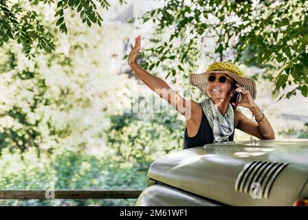 Eine Frau mit Hut und Sonnenbrille hebt ihre Hand in der Begrüßung und spricht am Telefon vor einem Geländewagen. Im Hintergrund ein Wald. Stockfoto