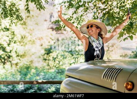 Eine Frau mit Hut und Sonnenbrille hebt die Arme vor einem Geländewagen. Im Hintergrund ein Wald. Konzeptnatur und Pkw-Routen. Stockfoto