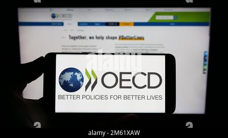 Person, die ein Mobiltelefon mit dem Logo der zwischenstaatlichen Organisation OECD auf dem Bildschirm vor der Webseite hält. Konzentrieren Sie sich auf das Display des Telefons. Stockfoto