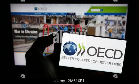 Person, die ein Mobiltelefon mit dem Logo der zwischenstaatlichen Organisation OECD auf dem Bildschirm vor der Webseite hält. Konzentrieren Sie sich auf das Display des Telefons. Stockfoto