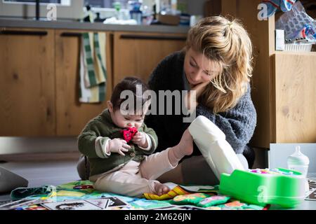 Die Mutter, die morgens mit ihrem Baby auf dem Boden in der Küche des chaotischen Hauses spielt, denkt darüber nach, was sie an diesem Tag tun muss. Stockfoto