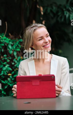 Frau, die wegsieht und lächelt, während sie ein digitales Tablet benutzt und draußen auf einem Tisch sitzt. Stockfoto