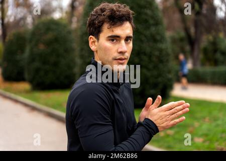 Seriöser junger Sportler mit kurzen Locken im schwarzen Rollkragenpullover, der sich auf der Straße im Park aufwärmt, während er in die Kamera schaut Stockfoto