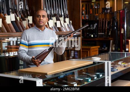 Gutaussehender, positiver erwachsener Mann im Jagdgeschäft mit seltenem Gewehr in den Händen Stockfoto