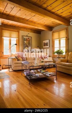 Cremefarbene und senffarbene Ledersofas und Glastisch im Familienzimmer mit lackierten Dielen in 1977 gebautem Blockhaus. Stockfoto