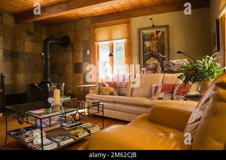 Cremefarbene und senffarbene Ledersofas und Glastisch im Familienzimmer mit schwarzem Gusseisenofen in 1977 gebautem Blockhaus. Stockfoto