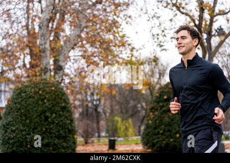 Seitenansicht des Content Guys in schwarzer Sportbekleidung, der auf dem unscharfen Hintergrund des Green Park joggt Stockfoto