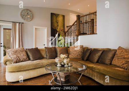 L-förmiges beige und braunes gepolstertes Sofa und runder Glas-Couchtisch im Wohnzimmer im luxuriösen Haus. Stockfoto