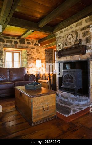 Braunes Ledersofa mit hölzernem Kofferraum-Couchtisch und Holzofen im Kamin im Wohnzimmer im alten, ca. 1850 Jahre alten Feldsteinhaus. Stockfoto