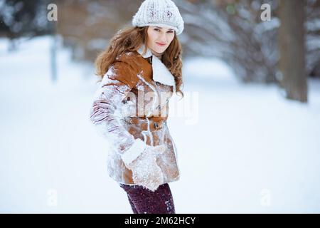Unzufriedene elegante 40-jährige Frau mit braunem Hut und Schal in Schaffellack, mit Fäustlingen, die im Winter in der Stadt den Schnee von der Kleidung schütteln. Stockfoto