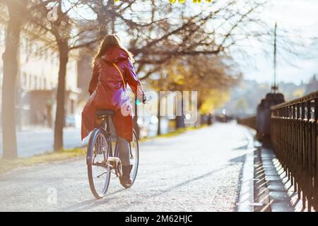 Von hinten gesehen Frau im roten Regenmantel im Freien in der Stadt Fahrrad fahren. Stockfoto