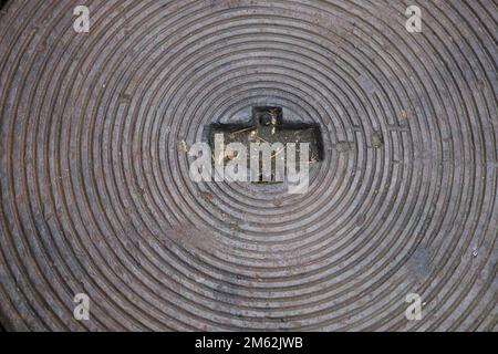 Gehweg, metallische Abdeckkappe mit konzentrischen Kreisen, die als strukturierter Hintergrund dient. In der Mitte befindet sich die Form eines Pluszeichens. Stockfoto