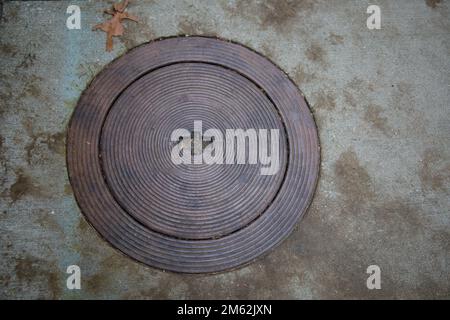 Einheimischer, schäbiger Bürgersteig einer Kleinstadt mit altmodischer, metallischer Haube mit konzentrischen Kreisen. Im oberen linken Bereich befindet sich ein getrocknetes Blatt. Stockfoto