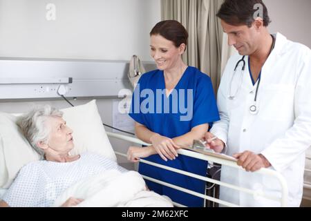 Deine Genesung läuft gut. Ein Arzt und seine Krankenschwester stehen über ihrem Patientenbett, während sie ein medizinisches Formular ausfüllen. Stockfoto